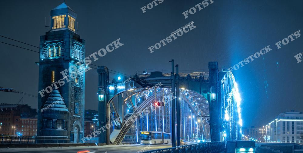 На фотографии изображен Большеохтинский мост в Санкт-Петербурге, освещенный холодными огнями зимнего вечера. Его массивные башни и металлические арки выделяются на фоне темного неба, подчеркивая индустриальную эстетику и силу конструкции. Этот мост, один из знаковых для города, соединяет берега Невы и привлекает внимание как своей архитектурой, так и инженерными особенностями. Легкий снегопад и мерцающий свет фар создают особую атмосферу зимней ночи в Петербурге.
